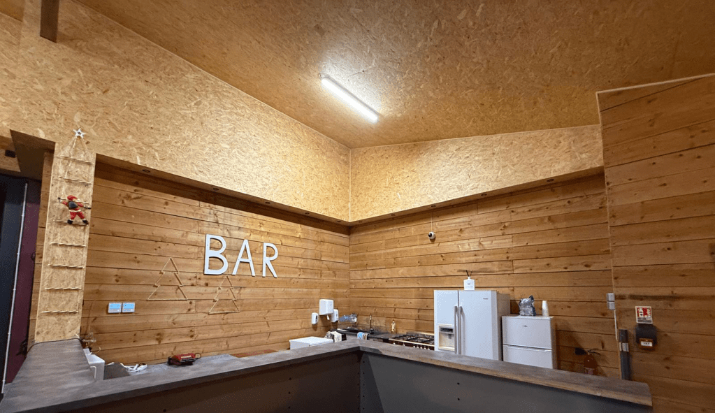 Intérieur d'un bar avec des murs en bois, une plaque 'BAR' en lettres blanches, un frigo et un espace de préparation, décoré d'une petite décoration de Noël.